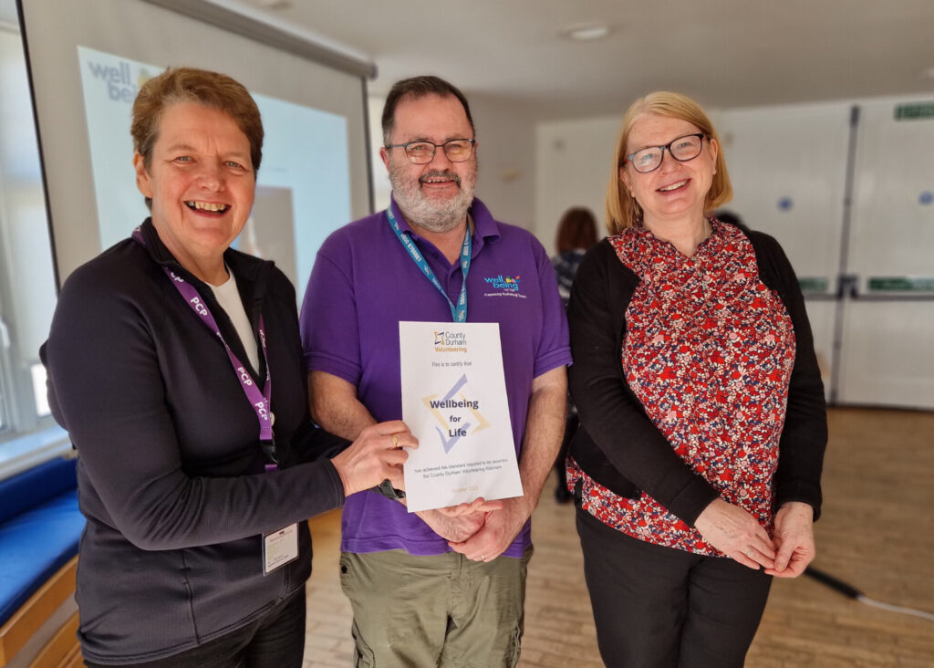 County Durham Wellbeing service recognised for quality volunteer experience 4 A group of 3 people. A woman with short hair is handing a certificate to a man in a Wellbeing for life uniform stood in the middle. A lady with a flowery blouse is stood next to them.