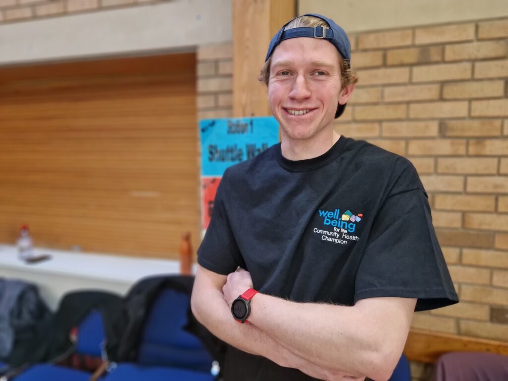 Durham University student adds value by volunteering 3 a young man stands, arms folded in a sports hall wearing a Wellbeing Community Champion t-shirt