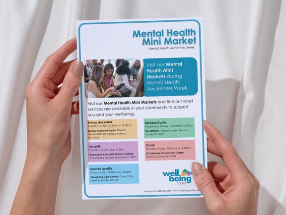 A close up of someone's hands holding a leaflet showing Wellbeing for Life's Mental Health Mini Market.
