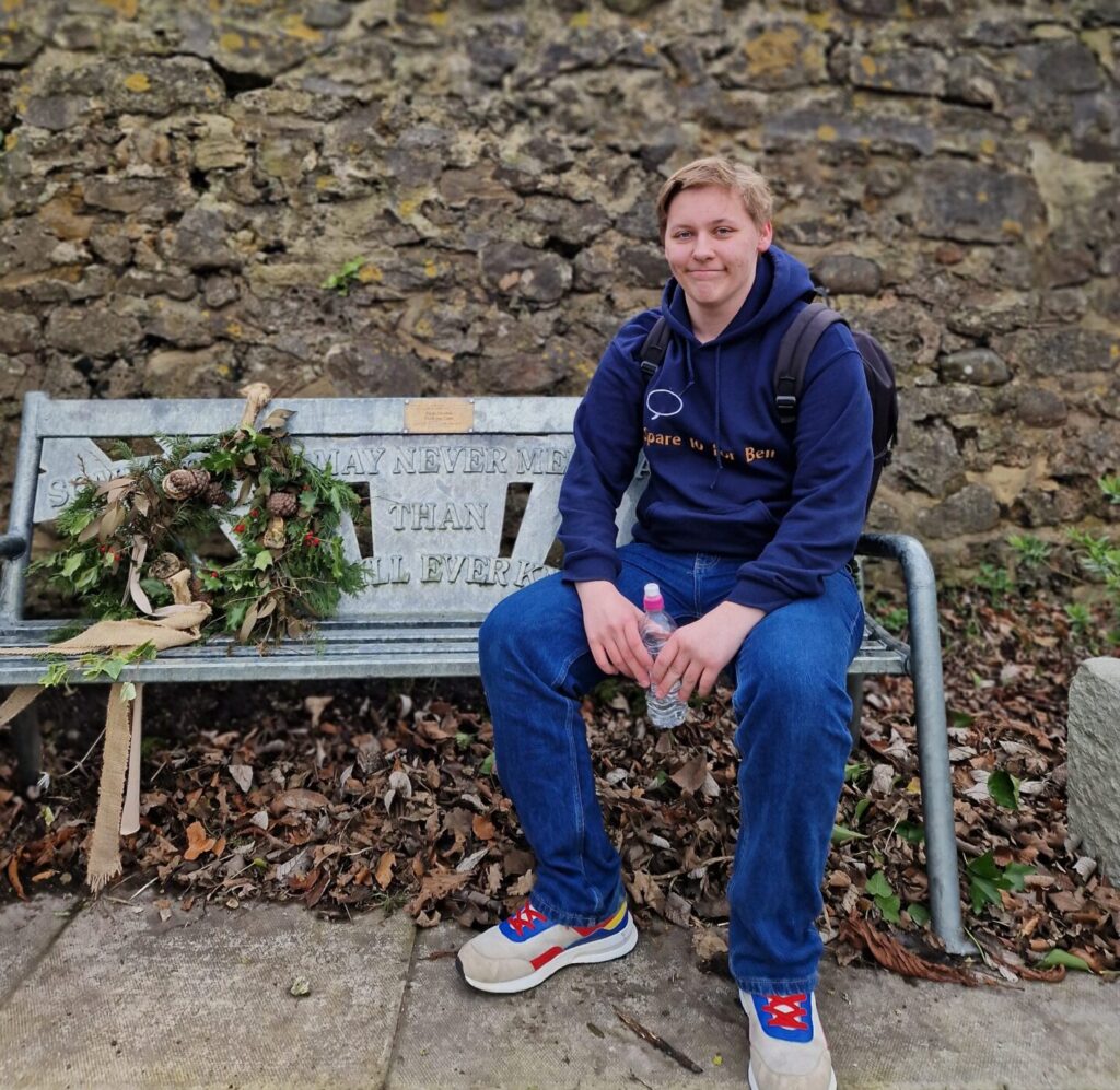 Volunteering at any age 2 A young male adult with short hair is sitting on a park bench wearing a blue hoody, a back pack and smiling