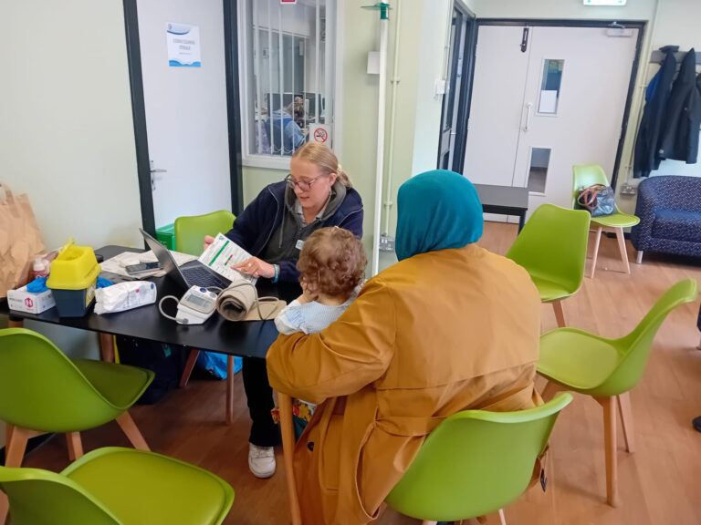 A lady wearing a head covering is sat to her back to the camera. There is a young child with curly hair sat on her knee. She is talking to a staff member from Health Squad who is pointing out information on a leaflet.