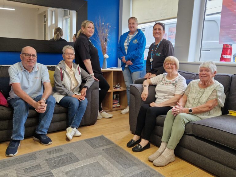 Two sofas are set at right angles and there are two people sat on each. In between the sofas are two members of Wellbeing for Life staff and a representative of The Cornforth Partnership