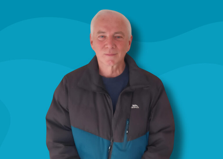 A man with grey hair is stood facing the camera. He is wearing a black and blue coat. The background is shades of blue in waves.