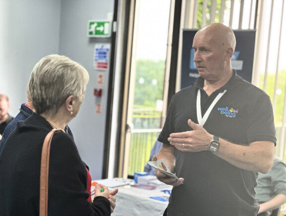 A tall man who works for Wellbeing for Life is chatting with a grey haired lady at an event.
