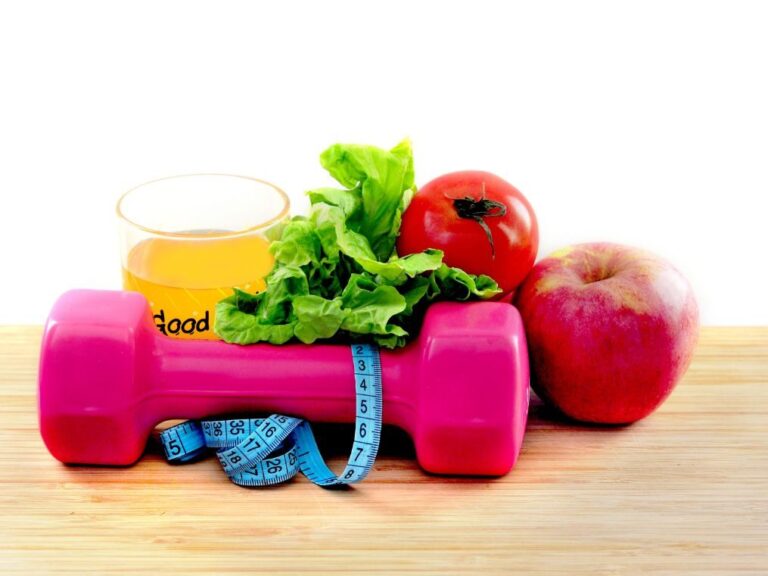 There are some items on a light wood table. In the forefront, there is a pink dumbbell with a tape measure draped over it. Behind it there is a cup of orange juice, a good helping of leafy lettuce, a tomato and an apple.