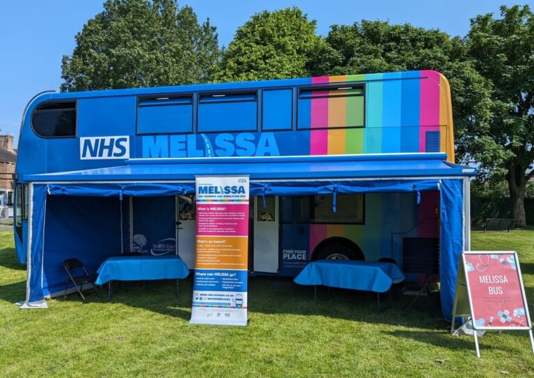 A picture of the Melissa Bus - A blue double decker bus with rainbow colours down the right hand side. In front of the bus there is a canopy, a banner and a sign about the melissa bus. and some table's and chairs underneath the canopy.