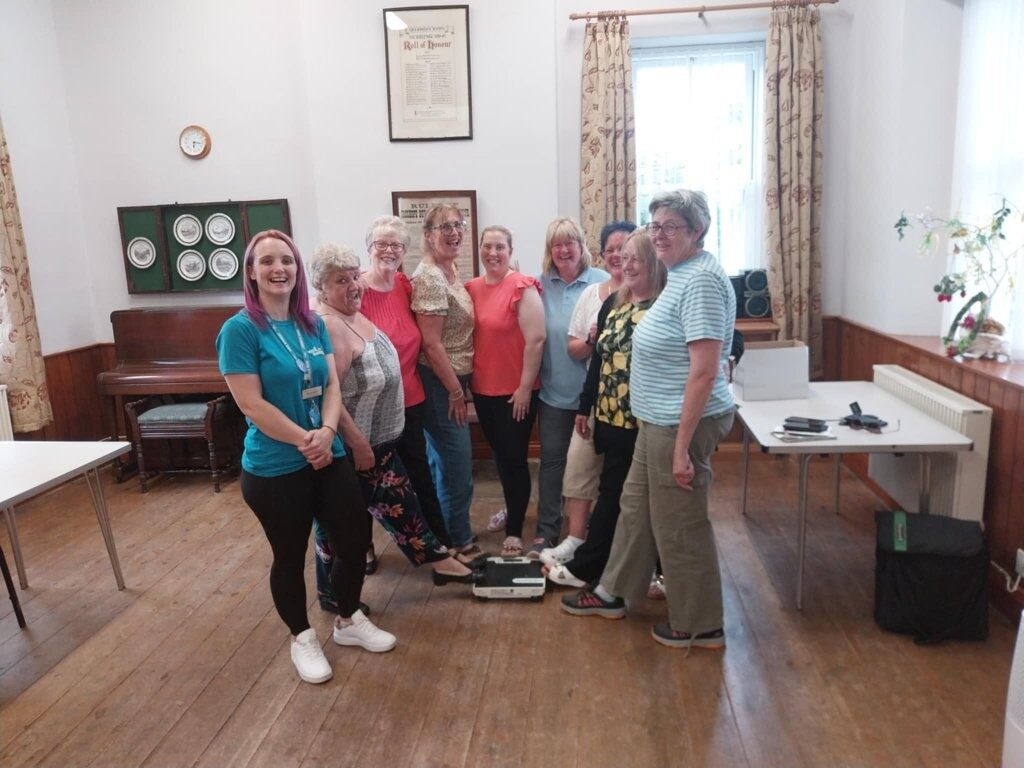 A group of 8 women standing together with big smiles on their faces. They all have 1 foot on some weight scales on the floor. Stood next to them is a Wellbeing for Life worker. She is also smiling.