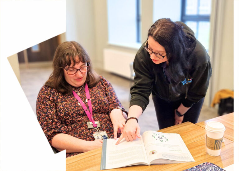 A trainer from Wellbeing for Life is chatting to a learner. They are pointing and a workbook and are engaging in conversation.