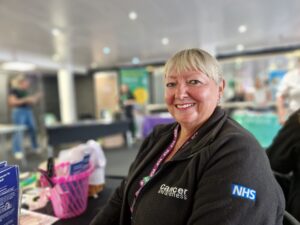 Claire is at a health and wellbeing event. Sat by her Cancer Awareness stall she is smiling and happy.