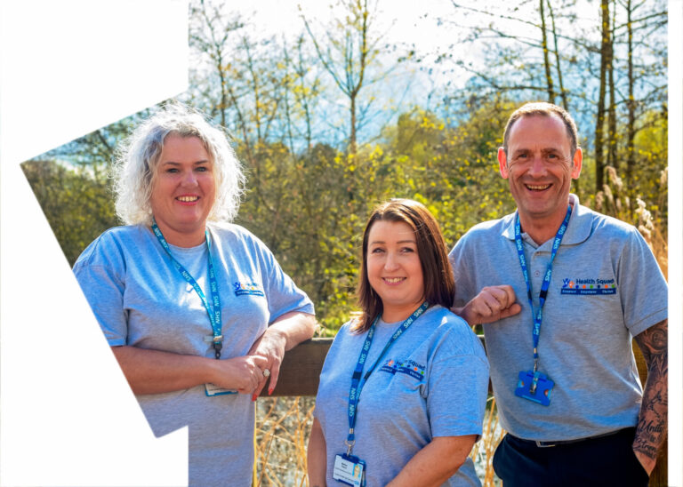 three members of the Health Squad team are stood together. Two are leaning on a fence rail - they are all smiling. There is trees and blue sky in the background