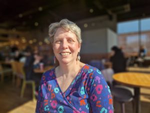 Lynne is sat in the sunshine and is smiling. She is wearing a blue dress with flowers on and her greying hair is cut in a short style.