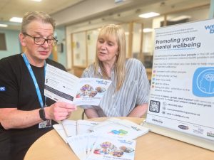 A male practitioner from Wellbeing for Life is chatting with a female client. They are looking at a service leaflet entitled, Your Wellbeing, Your Way. There are wellbeing workbooks and a roller banner on the table in front of them.