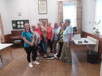 A group of 8 women standing together with big smiles on their faces. They all have 1 foot on some weight scales on the floor. Stood next to them is a Wellbeing for Life worker. She is also smiling.